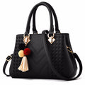 Black quilt-textured handbag with top handles, crossbody strap, and pom tassel charm