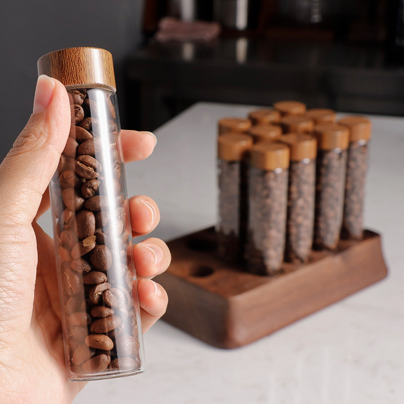 Hand holding a clear glass storage tube with cork top of coffee beans on a wooden stand